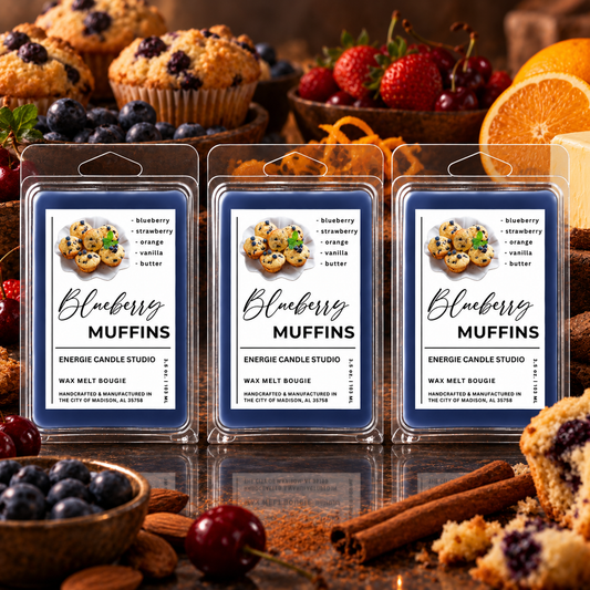 Three packaged wax melts labeled Blueberry Muffins arranged on a reflective surface, with muffins, berries, oranges, and cinnamon sticks used as styled props in a warm kitchen-like setting.