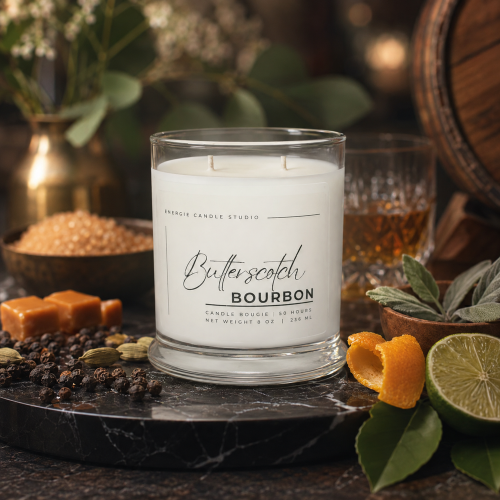 A white scented candle in a clear glass jar labeled 'Butter Scotch Bourbon' by Presque Candle Studio, styled on a dark marble surface with orange peels, a lime, whole spices, and a blurred whiskey glass and barrel in the background.