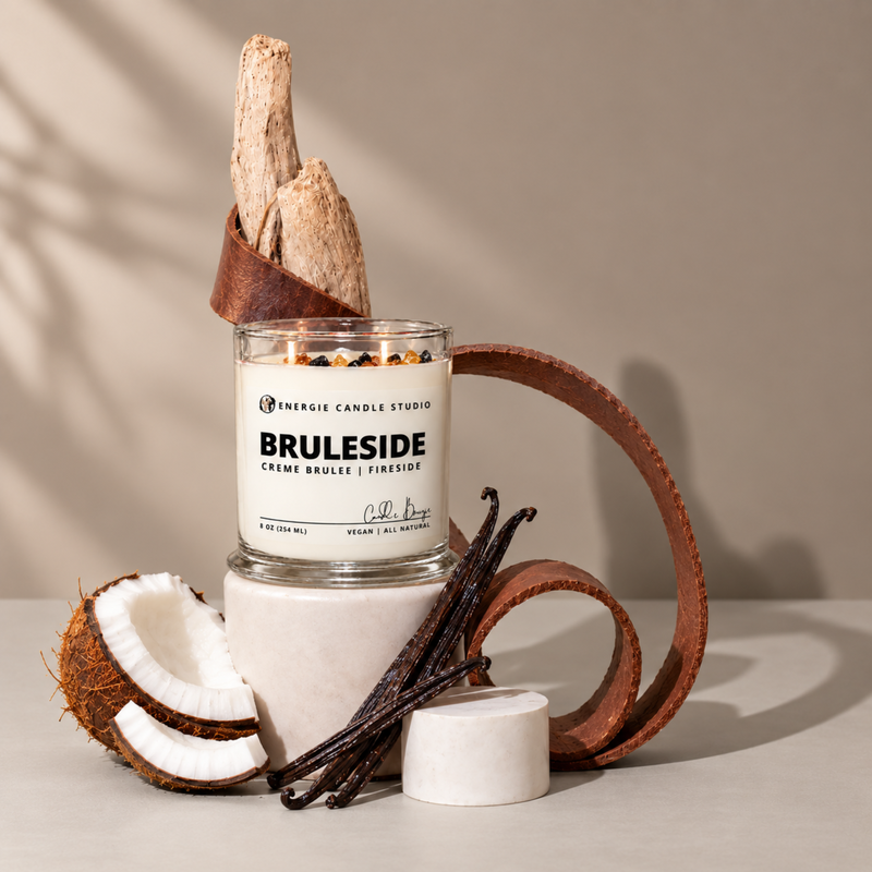 A styled product shot of a jar candle from Energie Candle Studio, labeled 'BRULESIDE'. The candle is set on a white marble pedestal with a brown leather loop, accompanied by a halved coconut, vanilla beans, and dried bark in a brown paper cone, all against a warm beige background.