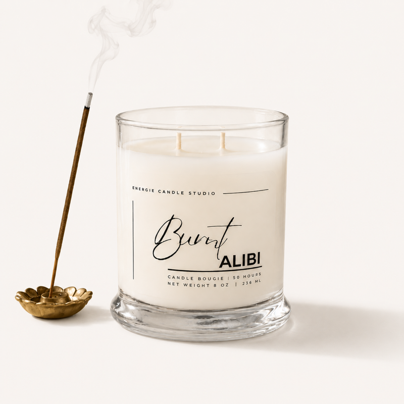 A clear glass jar candle with white wax sits on a light beige background, next to a gold incense holder with a smoking incense stick. The front label reads 'Burnt ALIBI' and 'ENERGIE CANDLE STUDIO'.