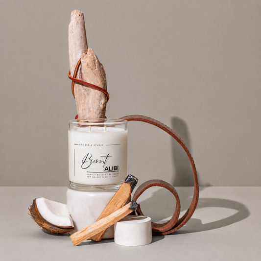 A glass jar candle labeled 'Breat ALIBI' by Smokey Candle Studio, styled with a driftwood piece wrapped in a leather cord, a coconut half, a cinnamon stick, and a small white container on a neutral studio background.