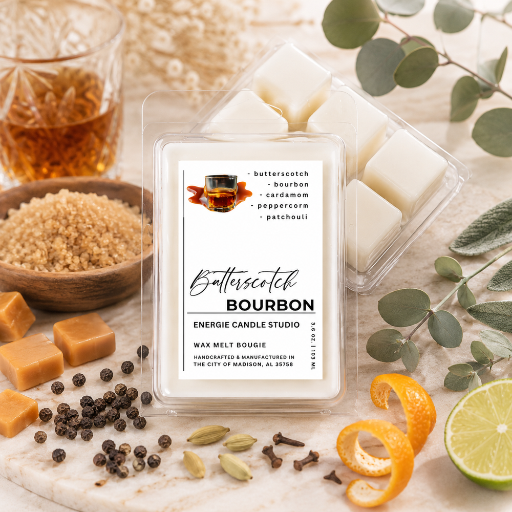 A wax melt package labeled 'Bitterscotch Bourbon' by Energie Candle Studio, shown in a clear plastic clamshell with white wax cubes visible. The package is styled on a marble surface with bourbon, spices, citrus, and eucalyptus leaves around it.
