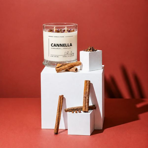 A glass jar candle labeled CANNELLA Cinnamon Scentile by Candle King sits on white display blocks with cinnamon sticks against a warm red background.