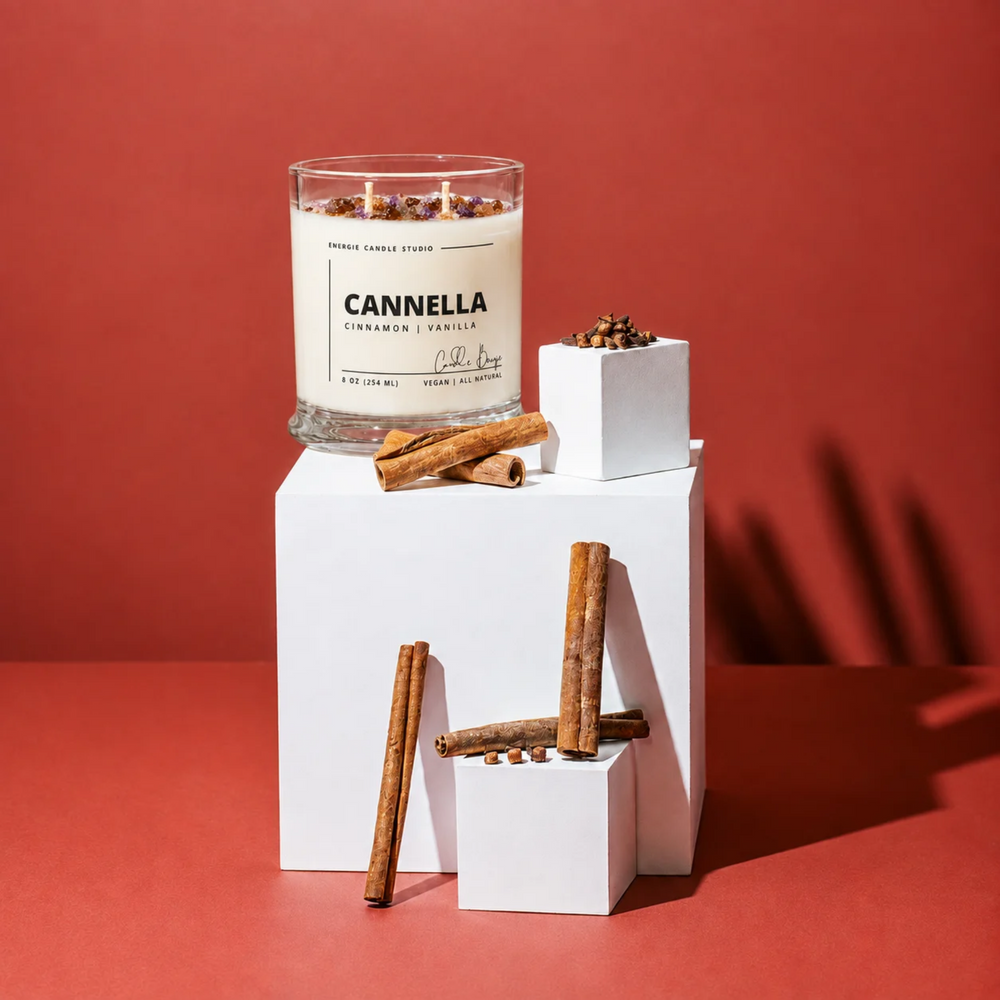 A glass jar candle labeled CANNELLA Cinnamon Scentile by Candle King sits on white display blocks with cinnamon sticks against a warm red background.