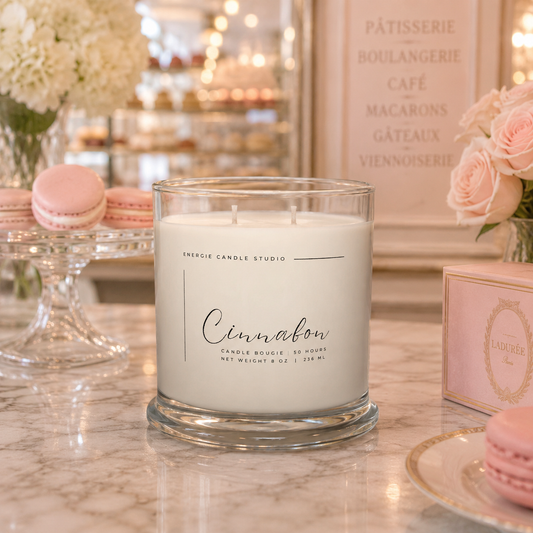 Candle labeled 'Cinnamon' on a marble surface with pastries and flowers in the background