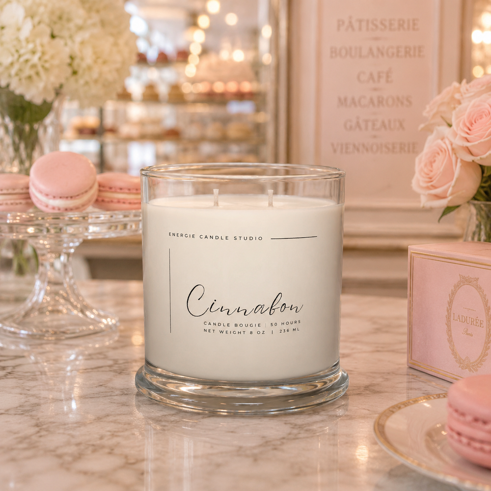 Candle labeled 'Cinnamon' on a marble surface with pastries and flowers in the background