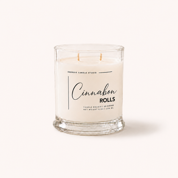 A double-wick candle in a clear glass jar with cream-colored wax, labeled 'Cinnabon Rolls' by Energy Candle Studio, set against a solid off-white background.