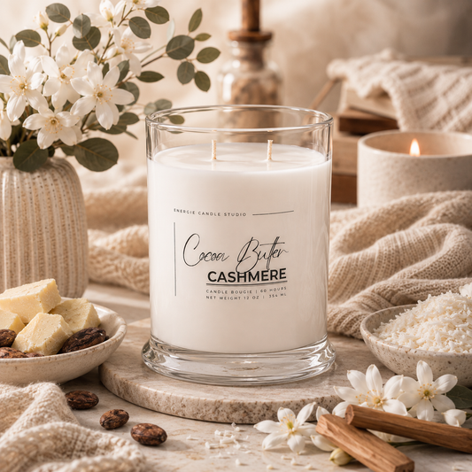 A double-wick candle in a clear glass jar labeled 'Cocoa Butter CASHMERE' by Candle Boutique Studio, styled on a stone board with cream cloth, flowers, cinnamon sticks, and small bowls of ingredients in a warm, cozy setting.