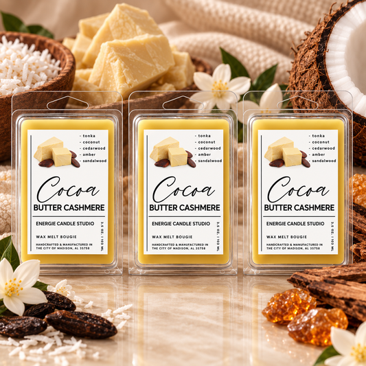 Three packaged wax melts labeled 'Cocoa Butter Cashmere' arranged in a styled product shot with cream-colored wax chunks, coconut halves, white flowers, and dried fruit on a reflective surface.