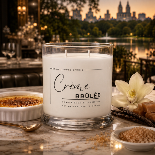 A white scented candle in a clear glass jar labeled 'Crème Brûlée' by Energie Candle Studio, styled on a marble surface with dessert dishes, a vanilla orchid, and a blurred city park background at sunset.