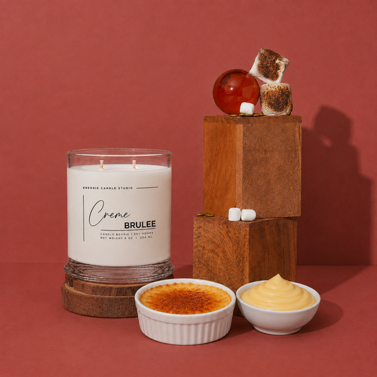A candle jar labeled 'Creme Brulee' by Breeze Candle Studio, styled with dessert props including a crème brûlée, a whipped cream bowl, marshmallows, and toasted marshmallows on wooden blocks against a terracotta background.