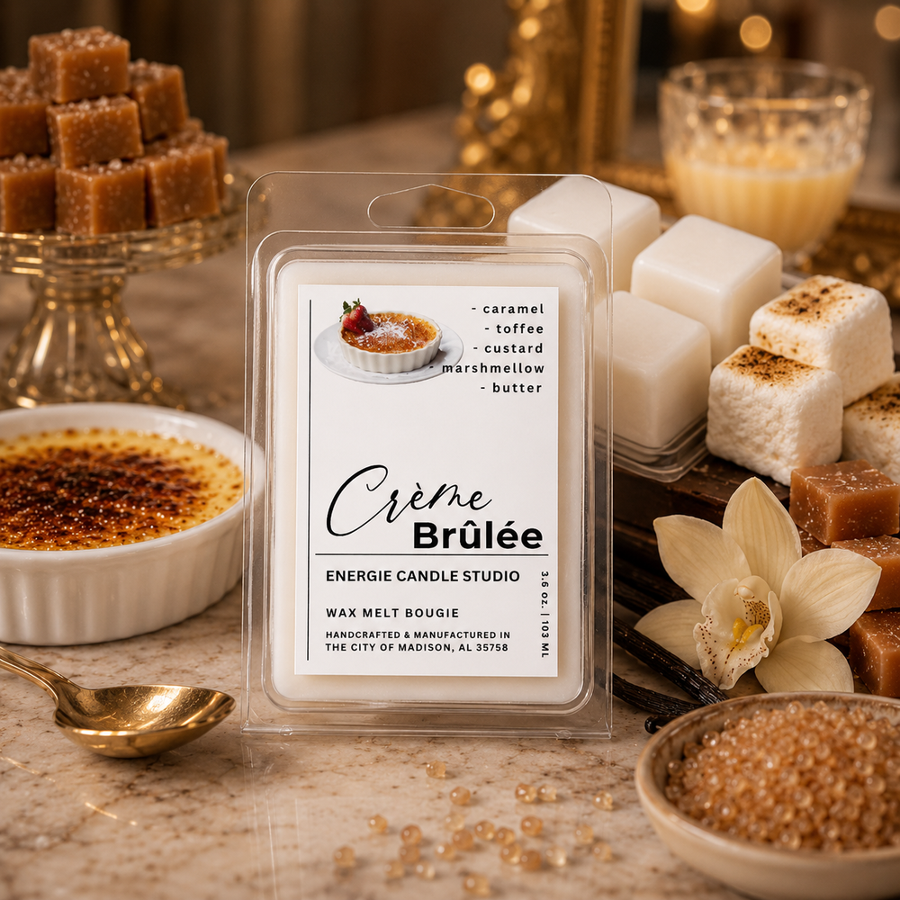 A clear plastic clamshell package labeled 'Crème Brûlée' wax melt is centered on a marble surface, surrounded by dessert-themed props including caramel brownies, marshmallows, a crème brulee dish, and a white orchid. The package text reads 'caramel - toffee - custard - marshmallow - butter' and shows the brand Energie Candle Studio.