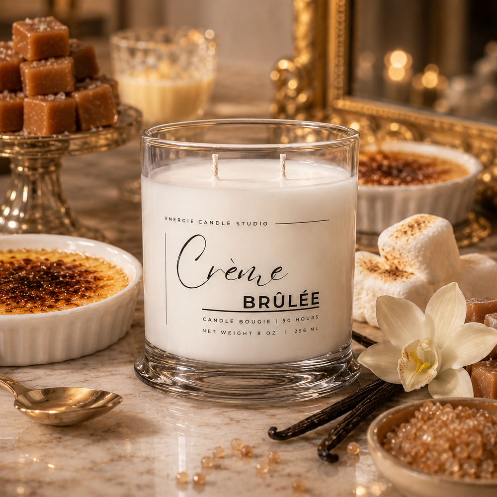 A white scented candle in a clear glass jar labeled “Crème Brûlée” by Energie Candle Studio, styled on a marble surface with caramel desserts, marshmallows, vanilla beans, and a white orchid flower.