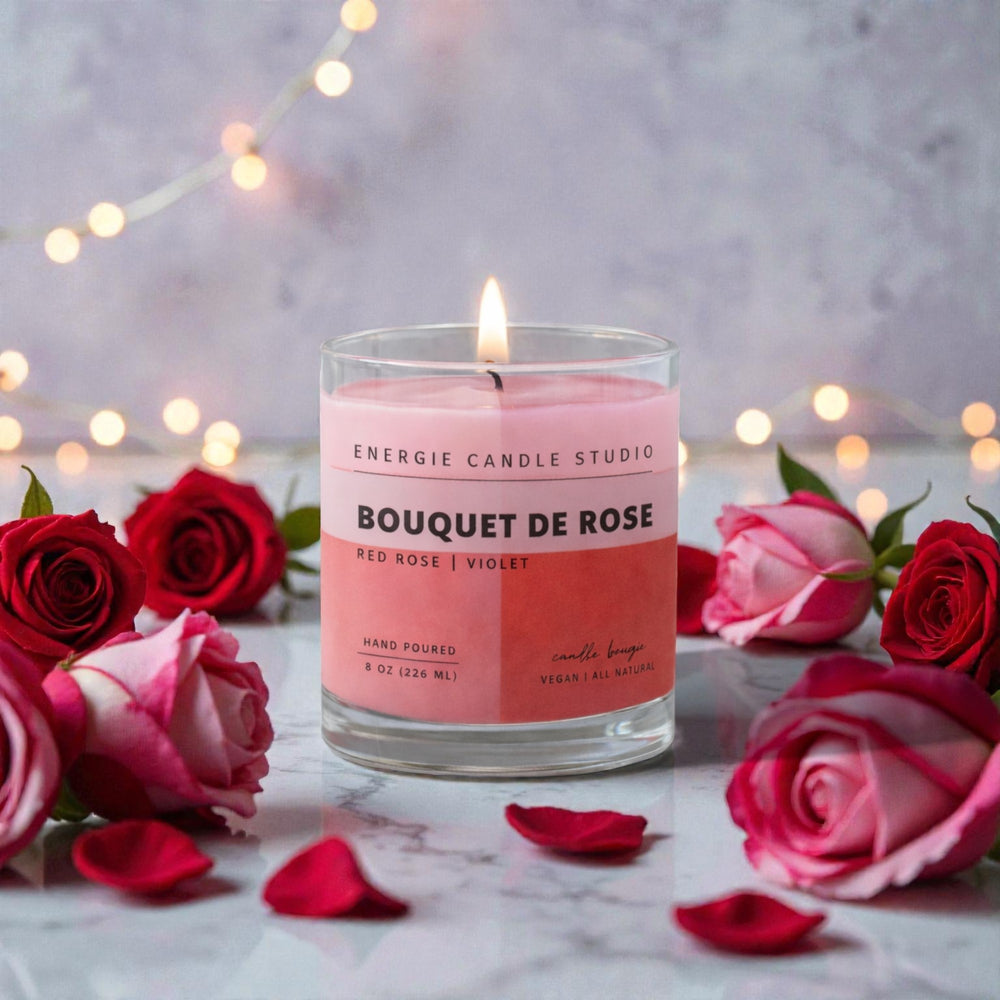 Pink candle labeled 'Bouquet de Rose' surrounded by red and pink roses on a marble surface with blurred lights in the background.