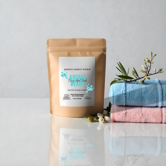 Beige packaging of Aroma Pearls with a white label, placed on a reflective surface with pink and blue towels and greenery in the background.