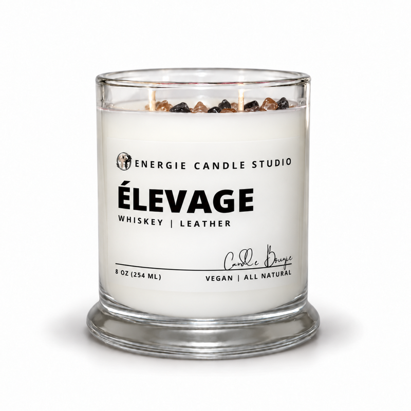 A 8 oz glass jar candle by Energie Candle Studio, featuring a white wax body, a clear glass container with a matching base, and decorative wood chips on top. The front label reads 'ÉLEVAGE WHISKEY | LEATHER' with a signature by Carl Beauge.