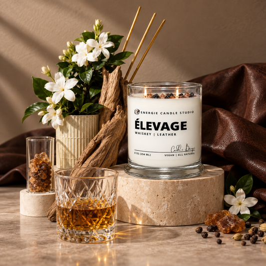 A lit scented candle in a clear glass jar sits on a stone pedestal in a styled setting with a whiskey glass, small condiment containers, white flowers, wood, and brown leather fabric. The candle label reads “ÉLEVAGE Whiskey | Leather” and shows the brand “Énergie Candle Studio.