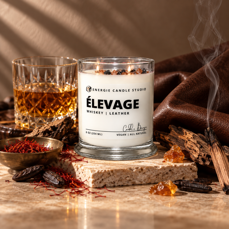 A lit candle in a clear glass jar labeled 'ÉLEVAGE Whiskey | Leather' by Energie Candle Studio, styled on a stone slab with whiskey, dried herbs, amber pieces, and a smoking incense stick on a warm brown background.