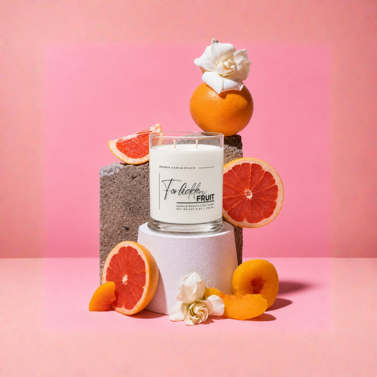 A double-wick scented candle in a clear glass jar sits on a white cylindrical pedestal, surrounded by grapefruit slices, orange peels, white flowers, and gray concrete blocks against a solid pink background. The candle label reads 'For the love of FRUIT' and 'ORANGE BLOSSOM SCENT'.