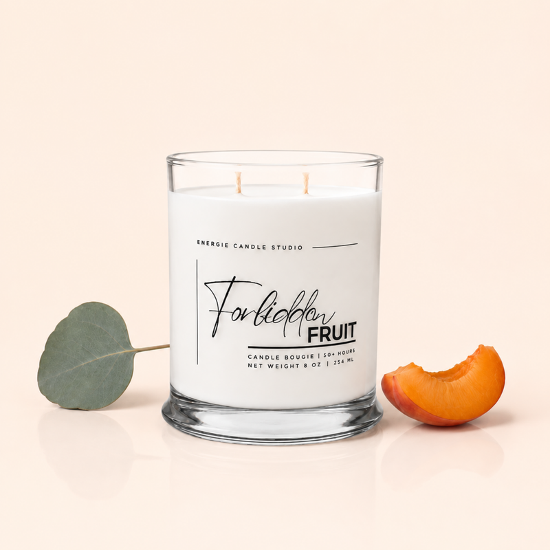A white scented candle in a clear glass jar with two wicks, labeled 'Forlóbble Fruit' by Energie Candle Studio, styled on a light beige background with a green eucalyptus leaf on the left and an apricot slice on the right.