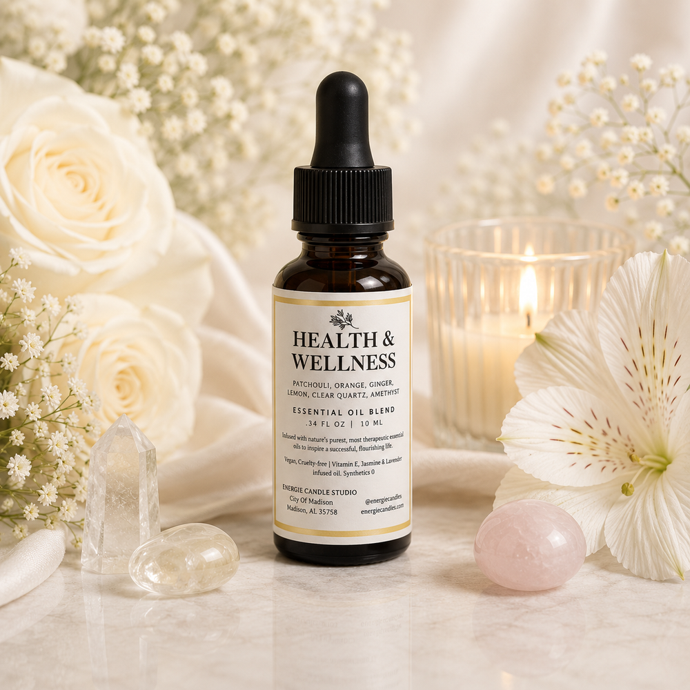 A small amber glass dropper bottle labeled Health & Wellness Essential Oil Blend stands on a light marble surface, surrounded by white flowers, crystal balls, and a lit candle in a glass holder.