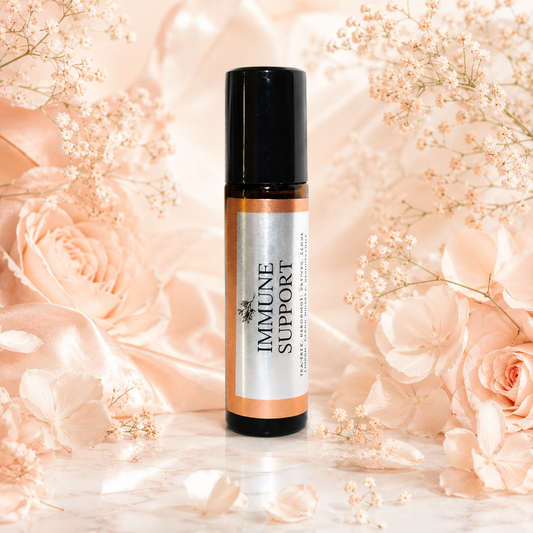 An amber glass roll-on bottle with a black cap and a silver label reading 'IMMUNE SUPPORT' is centered on a marble surface surrounded by soft peach satin, dried flowers, and rose petals.