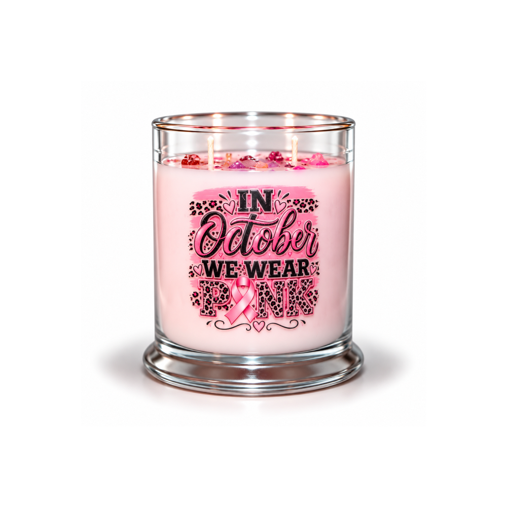A pink scented candle in a clear glass jar with a glass lid, featuring a graphic label that reads 'IN October WE WEAR PINK' with a pink ribbon and leopard print details. The candle is lit with three wicks, and the jar sits on a clean white background.