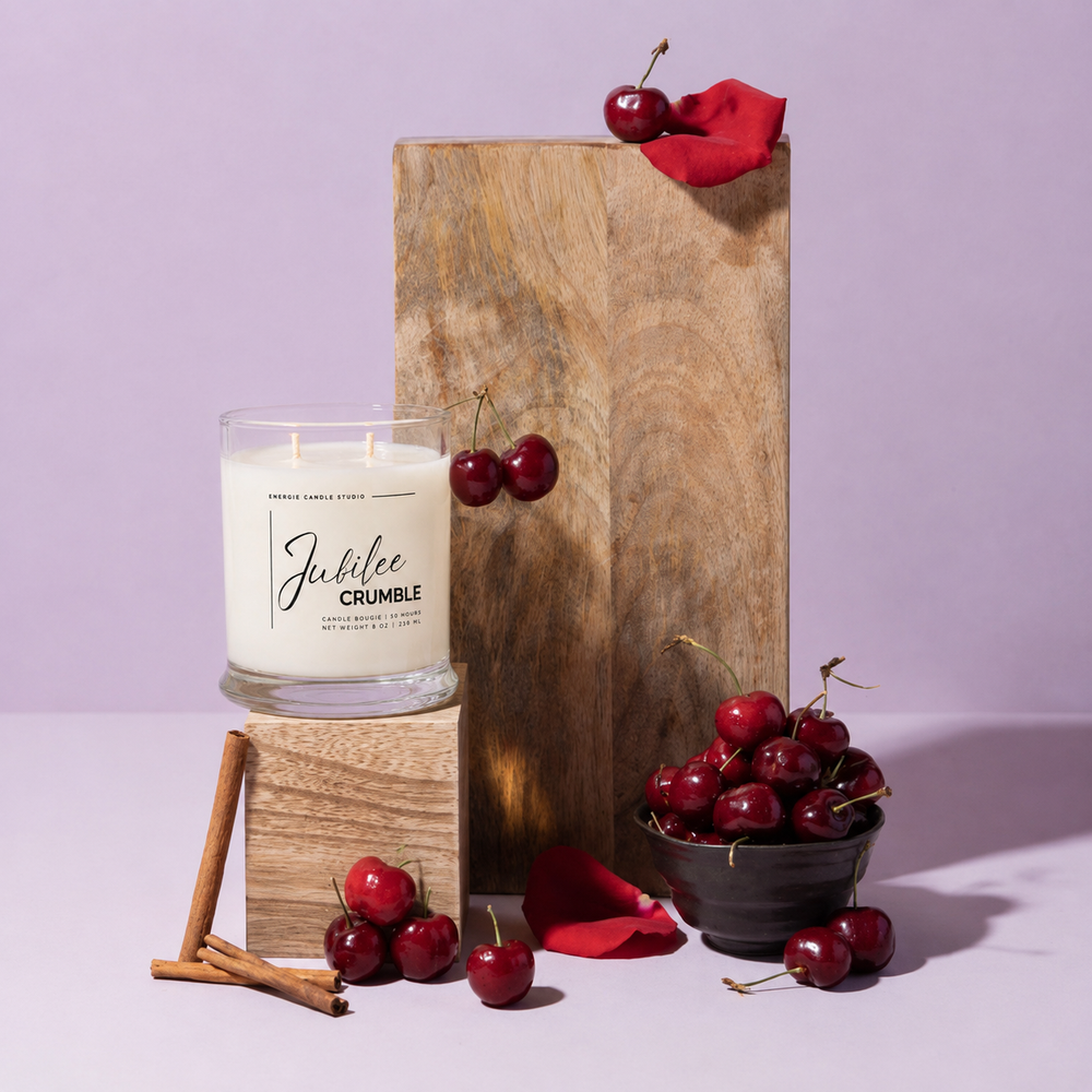 A lit scented candle in a clear glass jar sits on a wooden block, styled with fresh cherries, cinnamon sticks, and red rose petals around a tall wooden pedestal against a lavender background. The candle label reads 'Jubilee CRUMBLE' and 'Tennessee Cherry Wood'.