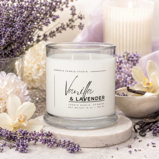 A glass jar candle labeled 'Vanilla & Lavender' by Energie Candle Studio, styled on a marble coaster with lavender flowers, white orchids, and vanilla beans in a soft, warm setting.