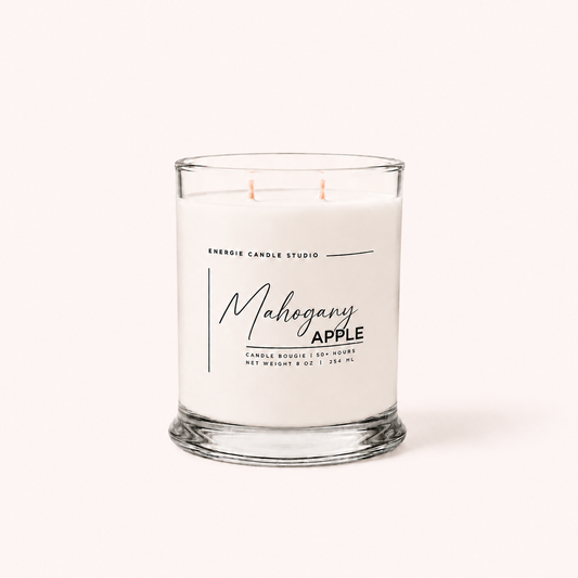 A white scented candle in a clear glass jar with two wicks, labeled 'Mahogany APPLE' by Energie Candle Studio, on a light cream background.