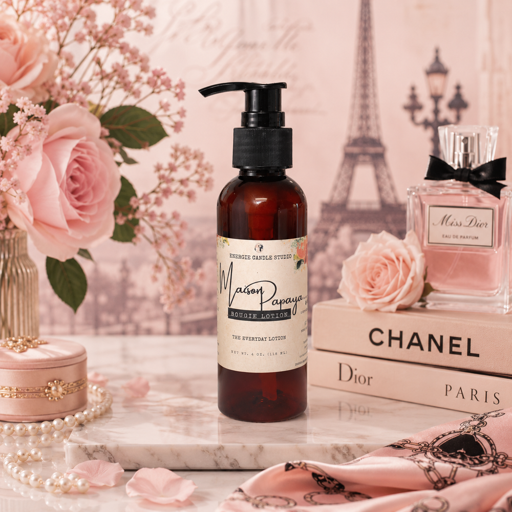 A bottle of Maison Papillon Sugar Scrub Lotion stands on a marble tray surrounded by pink roses, a perfume bottle, Chanel and Dior books, and a pink patterned scarf, with a soft pink Eiffel Tower backdrop.