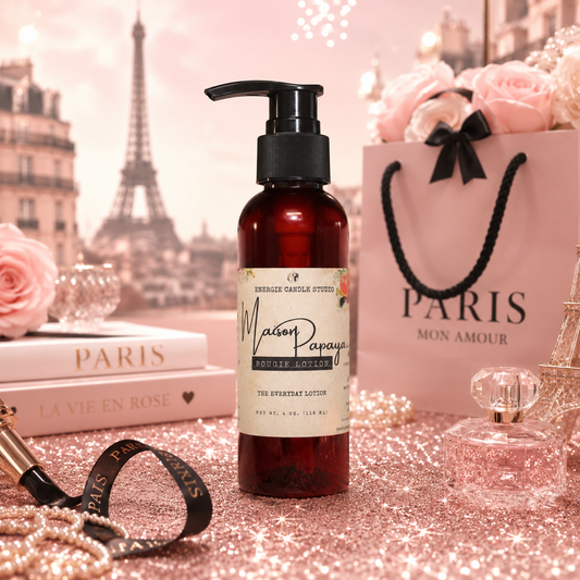 A bottle of Maison Papaya Exfoliating Lotion stands on a glittery pink surface with a Paris-themed background, including the Eiffel Tower, roses, a shopping bag, and perfume accessories.