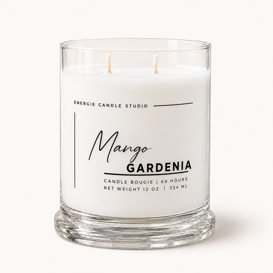 A two-wick scented candle in a clear glass jar with a thick base, featuring a white wax label with black text that reads 'Energie Candle Studio Mango Gardenia'.