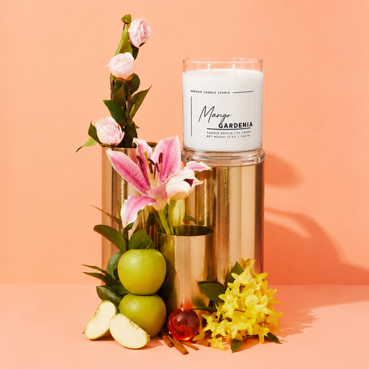 A glass jar candle labeled 'Mango Gardenia' by Freshly Picked Candle Studio, styled with pink flowers, green apples, and yellow blossoms in metallic gold cylindrical vases against a peach background.
