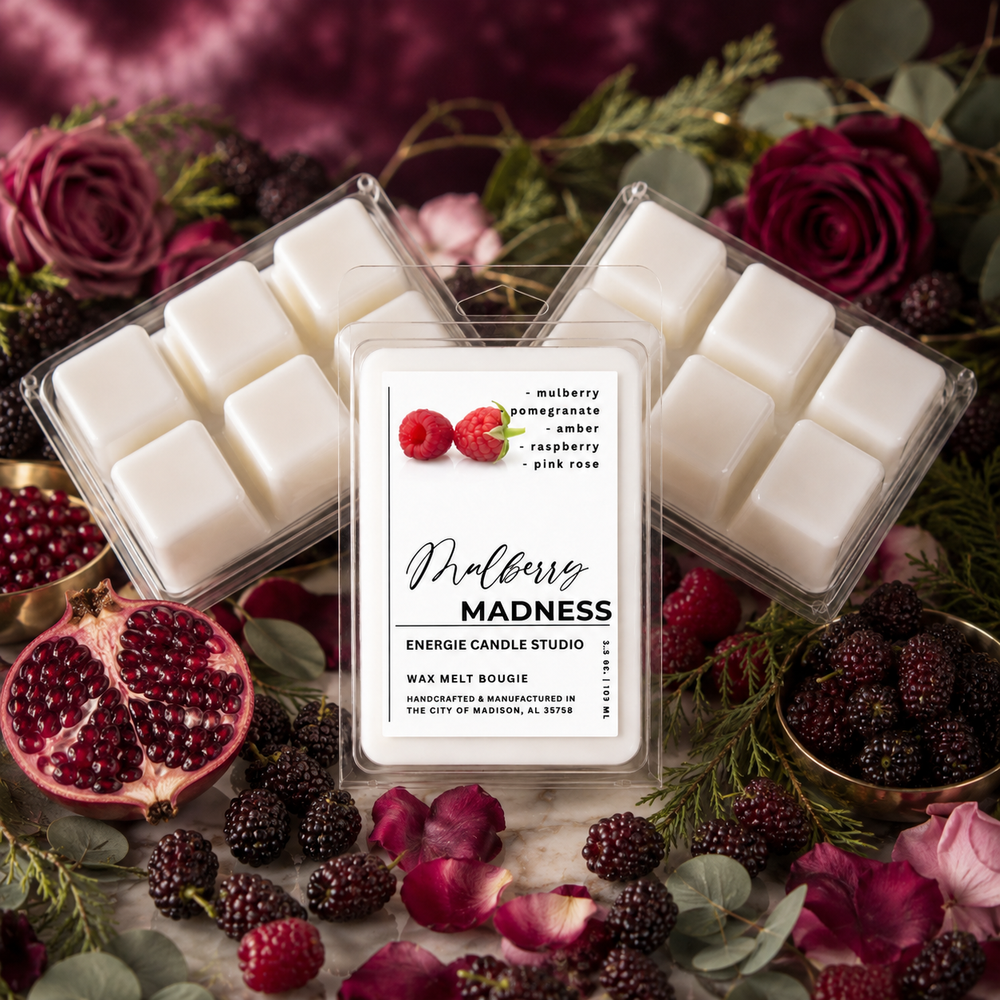 Three clear plastic wax melt packages filled with white wax melts are arranged on a marble surface surrounded by berries, pomegranate, roses, and greenery. The center package label reads 'Mulberry MADNESS' and shows berry illustrations.