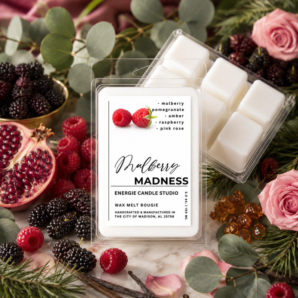 A clear plastic clamshell package of wax melts labeled 'Mulberry Madness' by Energie Candle Studio, surrounded by fresh berries, pomegranate, pink roses, eucalyptus leaves, and amber gummy bears on a marble surface.