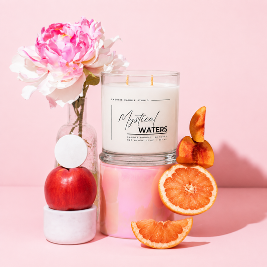 A glass jar candle labeled 'Mystical Waters' by Candle Candle Studio, styled on a pink surface with a pink flower in a glass vase, a red apple, and orange slices around it.