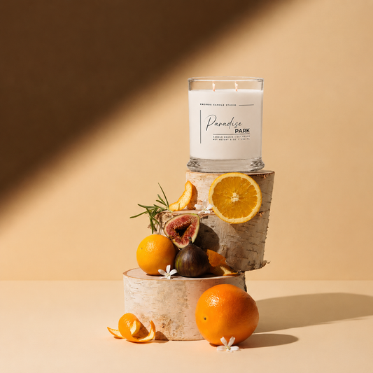 A white scented candle in a clear glass jar sits on stacked birch wood slices, styled with oranges, figs, orange slices, rosemary, and small white flowers against a warm beige background with diagonal shadows.
