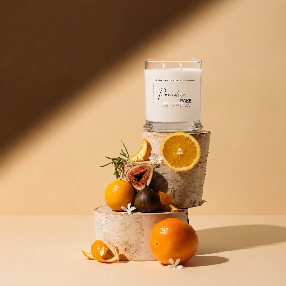 A white scented candle in a clear glass jar sits on stacked birch wood slices, styled with oranges, figs, orange slices, rosemary, and small white flowers against a warm beige background with diagonal shadows.