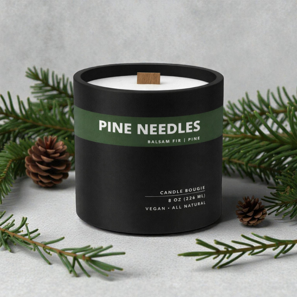 Candle labeled 'Pine Needles' with green label and black container, surrounded by pine branches and cones on a light gray background.