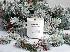 Candle labeled 'Pine Needles' surrounded by artificial Christmas trees with red berries on a light background