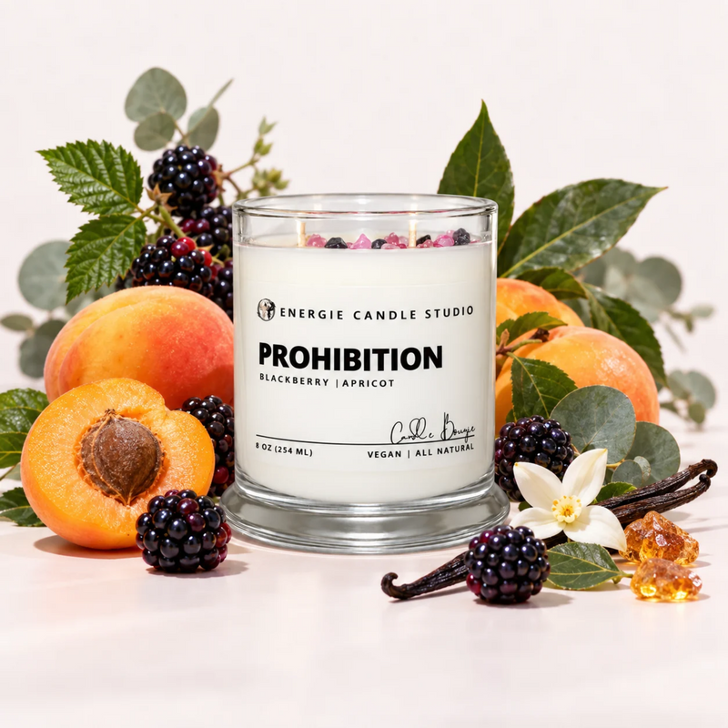A glass jar candle labeled 'PROHIBITION BLACKBERRY/APRICOT' by Energie Candle Studio, styled with apricots, blackberries, eucalyptus leaves, vanilla beans, and a white flower on a clean white background.