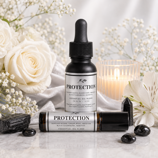 A small amber glass essential oil bottle with a black dropper cap stands behind a matching oil roller bottle labeled PROTECTION, styled on a light marble surface with white flowers, black crystals, and a lit candle in the background.