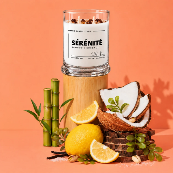 A glass jar candle labeled SÉRÉNITÉ Bamboo | Coconut by Evening Candle Studio, styled on a wooden pedestal with coconuts, lemons, bamboo, and greenery against a peach studio background.