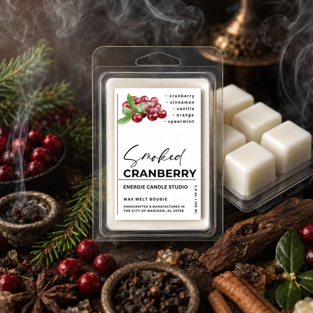 A packaged wax melt candle labeled 'Smoked Cranberry' by Energie Candle Studio, shown in a clear clamshell package with white wax cubes visible. The product is styled with cranberries, evergreen branches, a smoky mug, cinnamon sticks, and dark wood on a rustic surface.