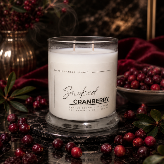 A double-wick candle in a clear glass jar labeled 'Smoked Cranberry' by Energie Candle Studio, styled with fresh cranberries on a dark marble surface and deep red fabric in the background.