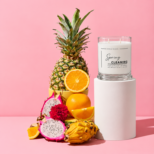 A scented candle in a clear glass jar sits on a white cylindrical pedestal against a pink studio background. The jar label reads 'Spring Cleaning' and 'Tropical Sandal Studio.