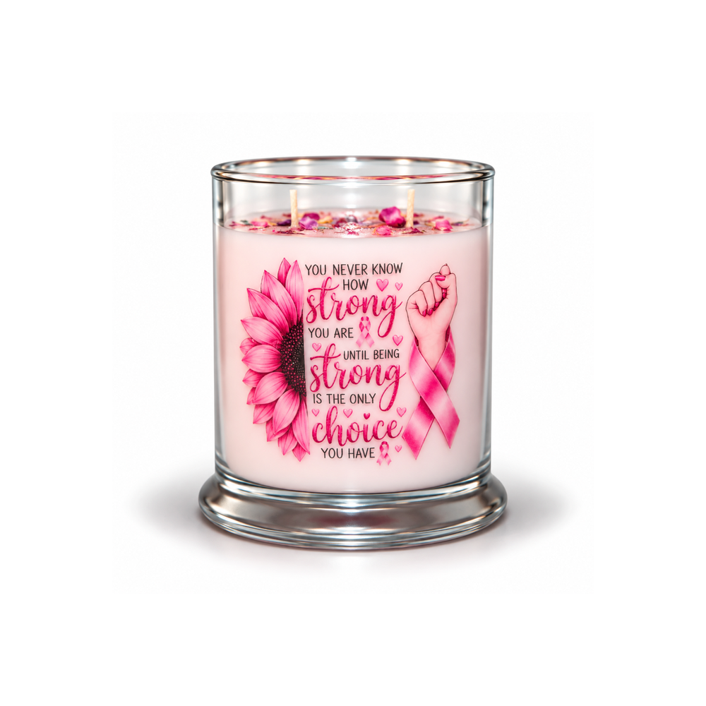 A pink scented candle in a clear glass jar with a silver-toned base, featuring a pink awareness ribbon, a pink sunflower, and the quote 'You never know how strong you are until being strong is the only choice you have'.