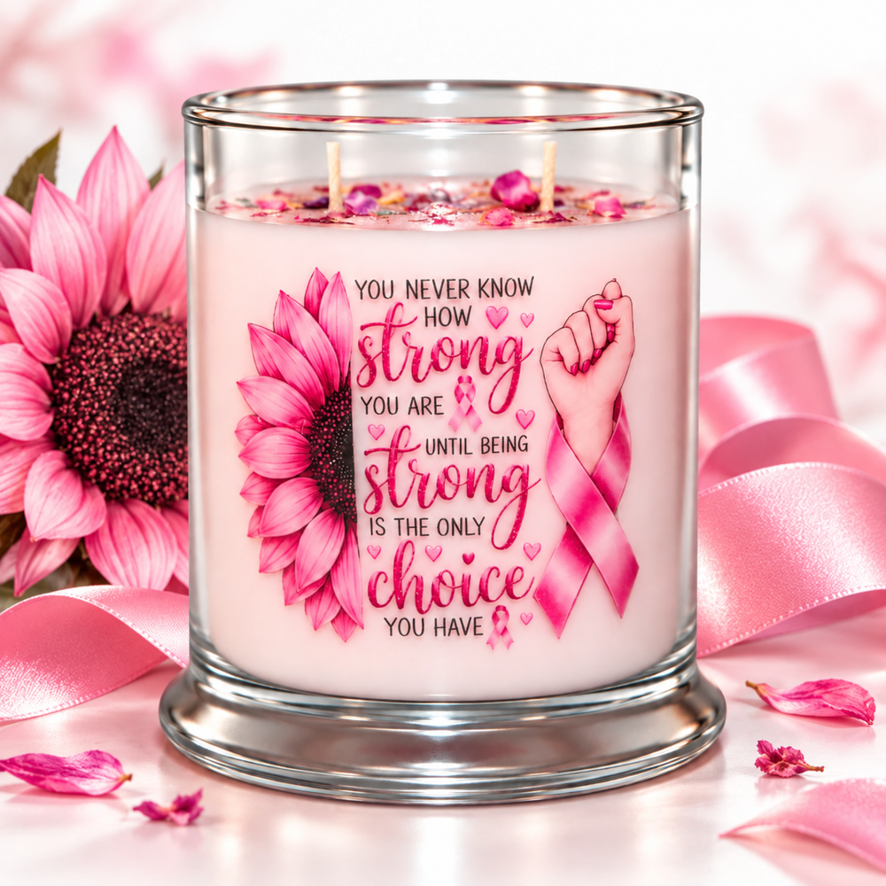 A pink candle in a clear glass jar with two wicks, decorated with a pink sunflower, a pink awareness ribbon, and a fist graphic with the text 'YOU NEVER KNOW HOW strong YOU ARE UNTIL BEING STRONG IS THE ONLY choice YOU HAVE.' The candle is styled with pink ribbons, sunflowers, and petals on a soft pink background.