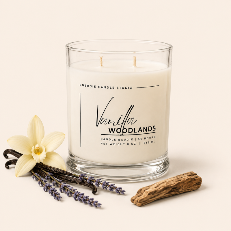 A two-wick scented candle in a clear glass jar by Energie Candle Studio, labeled Vanilla Woodlands. The candle is styled on a warm cream background with a vanilla flower, lavender sprigs, a vanilla bean, and a piece of driftwood.